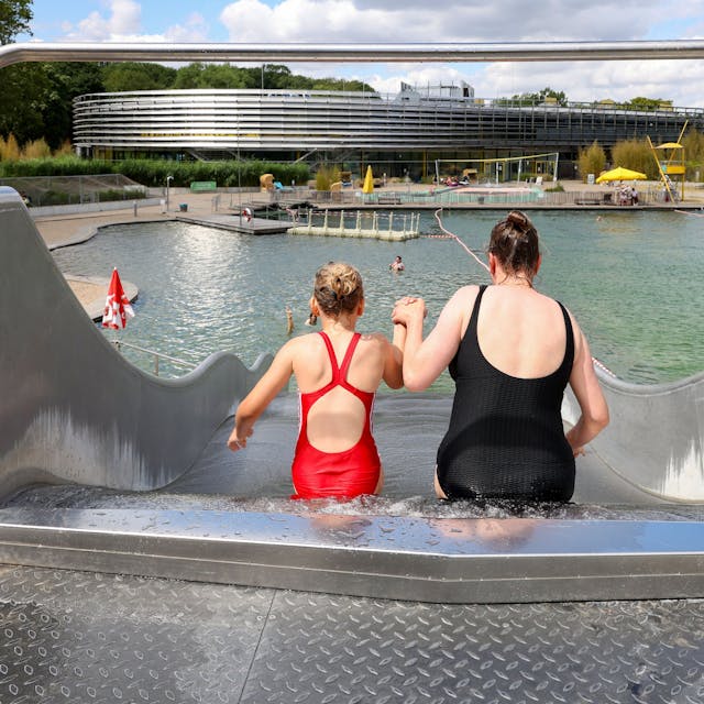 Das Bild zeigt eine Mutter und ihre Tochter von hinten, die einen Wasserrutsche hinunterrutschen. Im Hintergrund sieht man den Naturbadeteich im Lentpark Freibad in Köln.