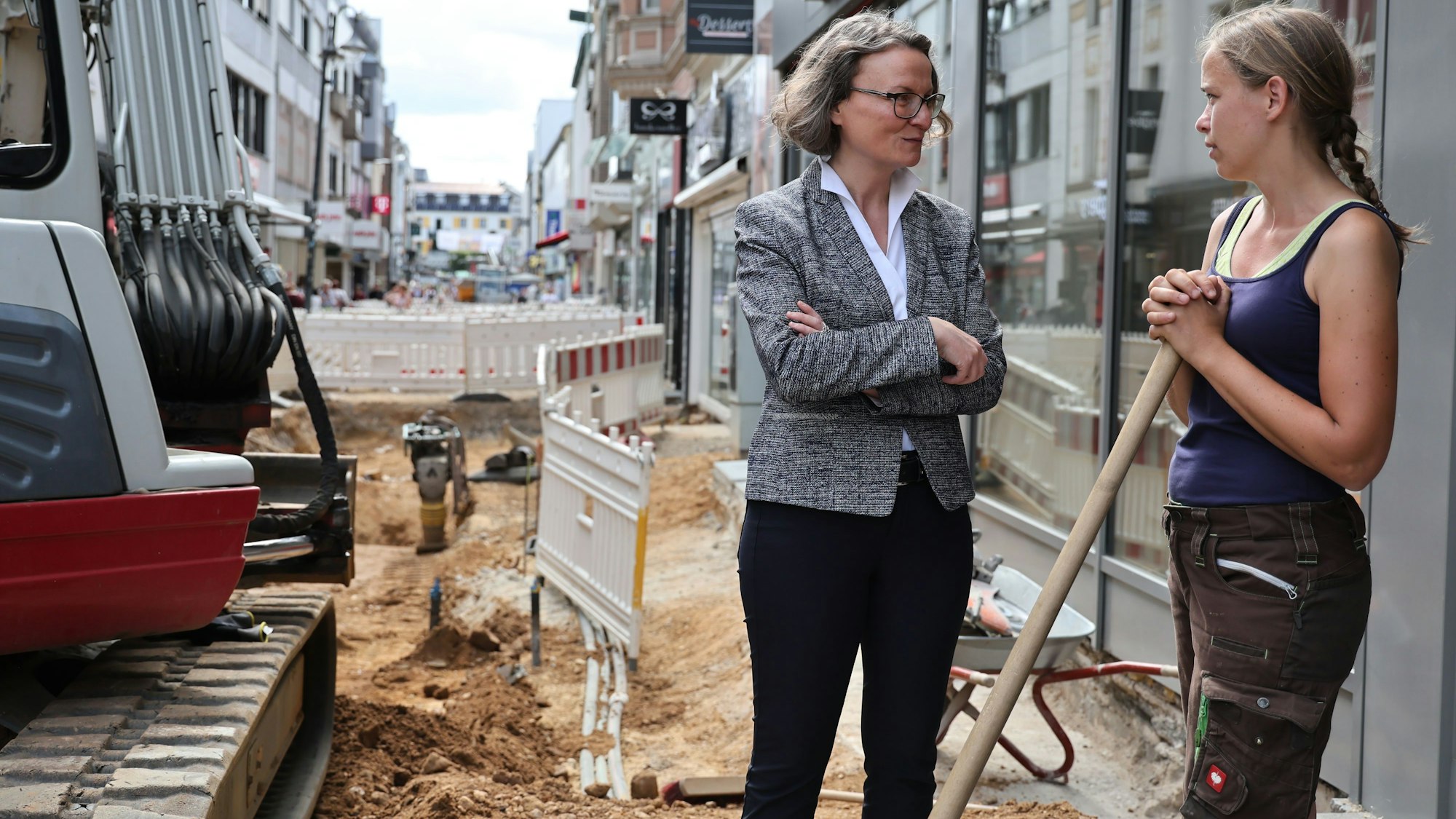 Die Euskirchener Innenstadt ist eine einzige Baustelle. Hier wird neues Pflaster verlegt. An der Baustelle, in der ein Kleinbagger steht, spricht Ina Scharrenbach mit der Praktikantin der Baufirma.