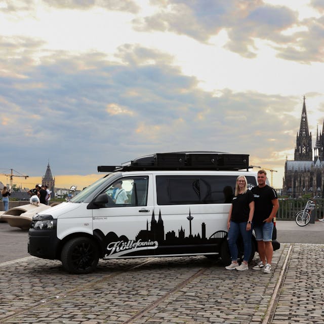 Das Foto zeigt den Bulli und Verena Van der Vight und Tim Locke vor der Aussichtsplattform in Köln. Im Hintergrund sieht man die Hohenzollernbrücke, den Kölner Dom und den Groß St. Martin.