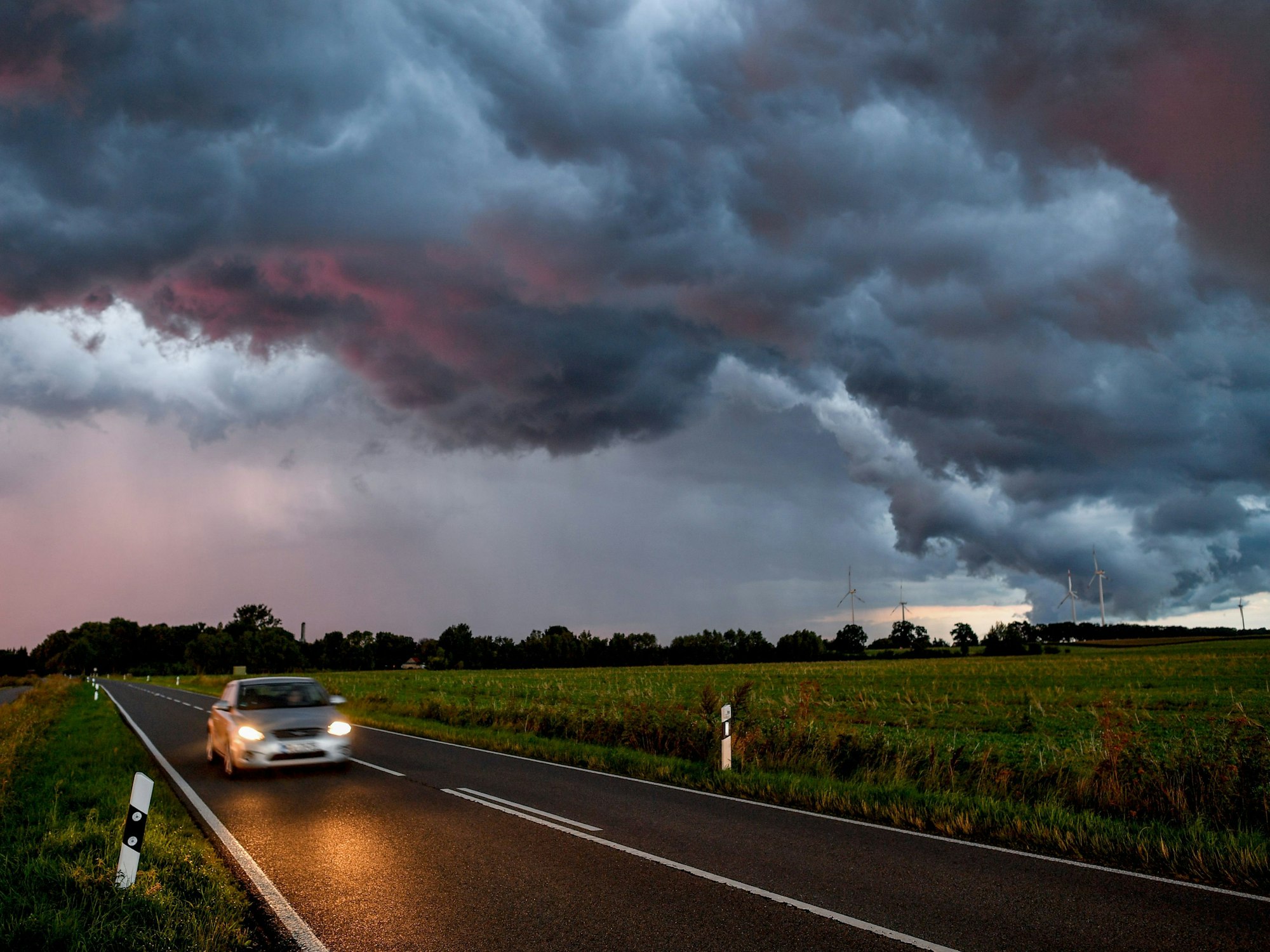 Ein Auto fährt auf einer Straße. Im Hintergrund nähert sich ein Unwetter mit Blitz und Donner.