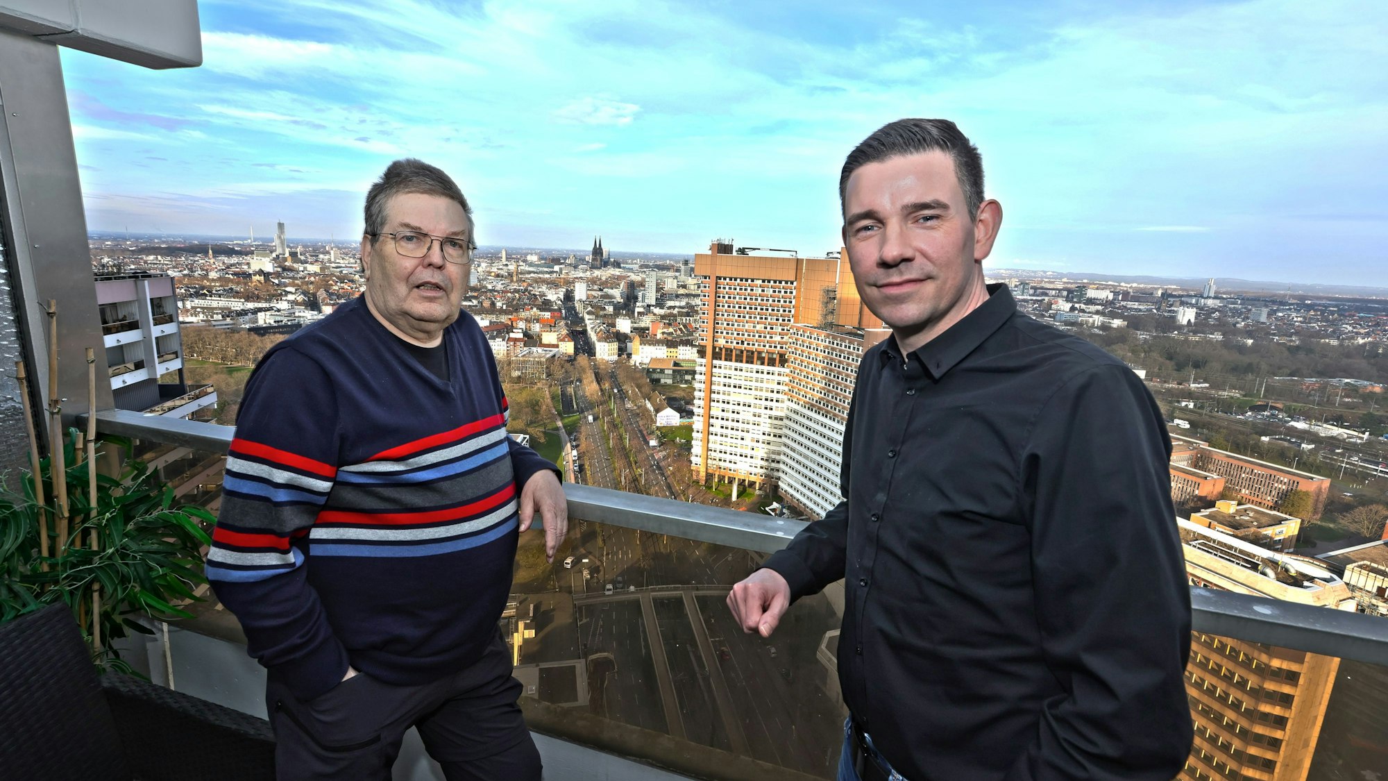 Zwei Männer auf einem Balkon, im Hintergrund sieht man die Skyline von Köln.