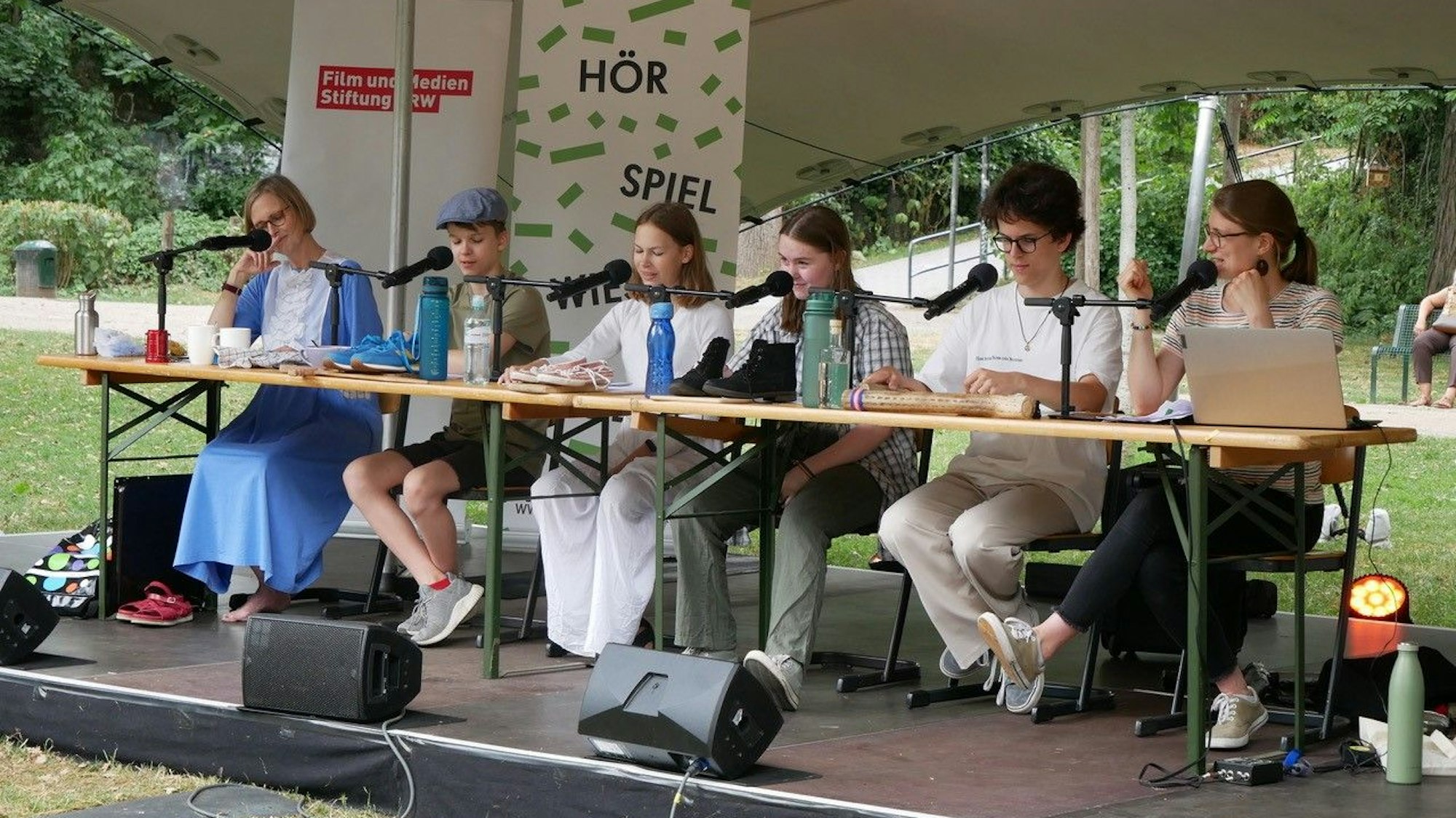 Zwei Erwachsene und vier Kinder sitzen auf einer Bühne im Park vor Mikrofonen.