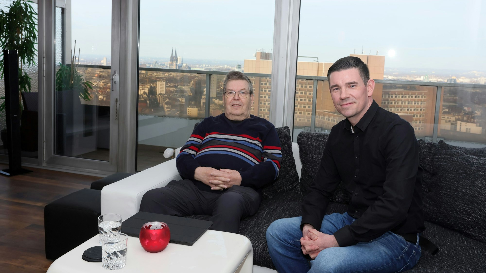 Zwei Männer sitzen auf einem Sofa, mit der Kölner Skyline im Hintergrund.
