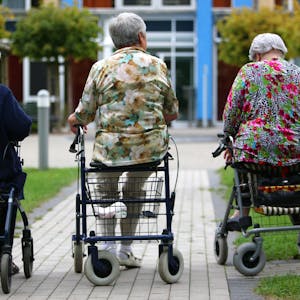 Drei Frauen höheren Alters sitzen auf Rollatoren auf einem Weg, der zu einem Pflegeheim führt