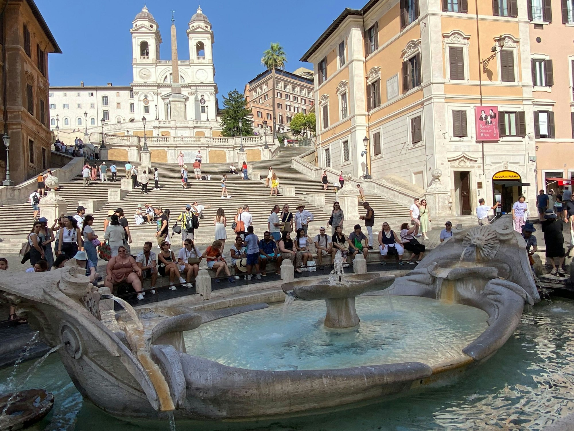 Touristinnen und Touristen sitzen im Schatten an der Fontana della Barcaccia, ein Brunnen auf der Piazza di Spagna, vor der Spanischen Treppe.