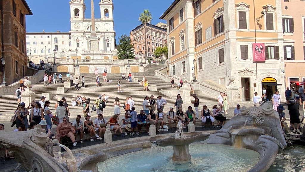 Touristinnen und Touristen sitzen im Schatten an der Fontana della Barcaccia, ein Brunnen auf der Piazza di Spagna, vor der Spanischen Treppe.