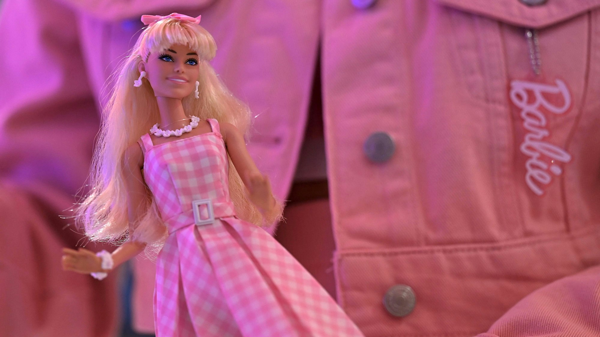 Barbie-Puppe vor einer rosa Jeansjacke mit Barbie-Logo