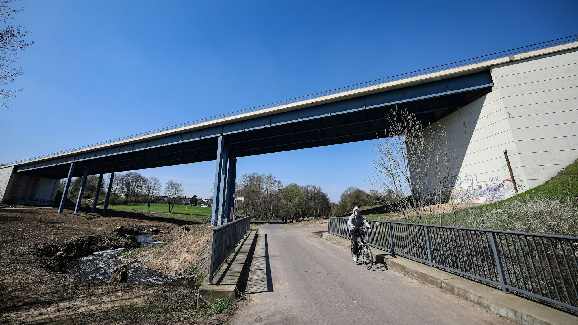 Ein seitlicher Blick auf die Haarbachtalbrücke in Aachen.Links ist ein Bach zu sehen, rechts fährt ein Radfahrer auf einem Weg.