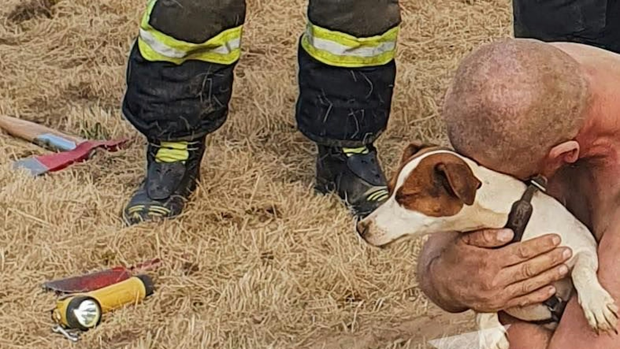 Ein Mann hockt auf dem Boden und drückt einen kleinen Hund an sich, Feuerwehrleute stehen daneben.