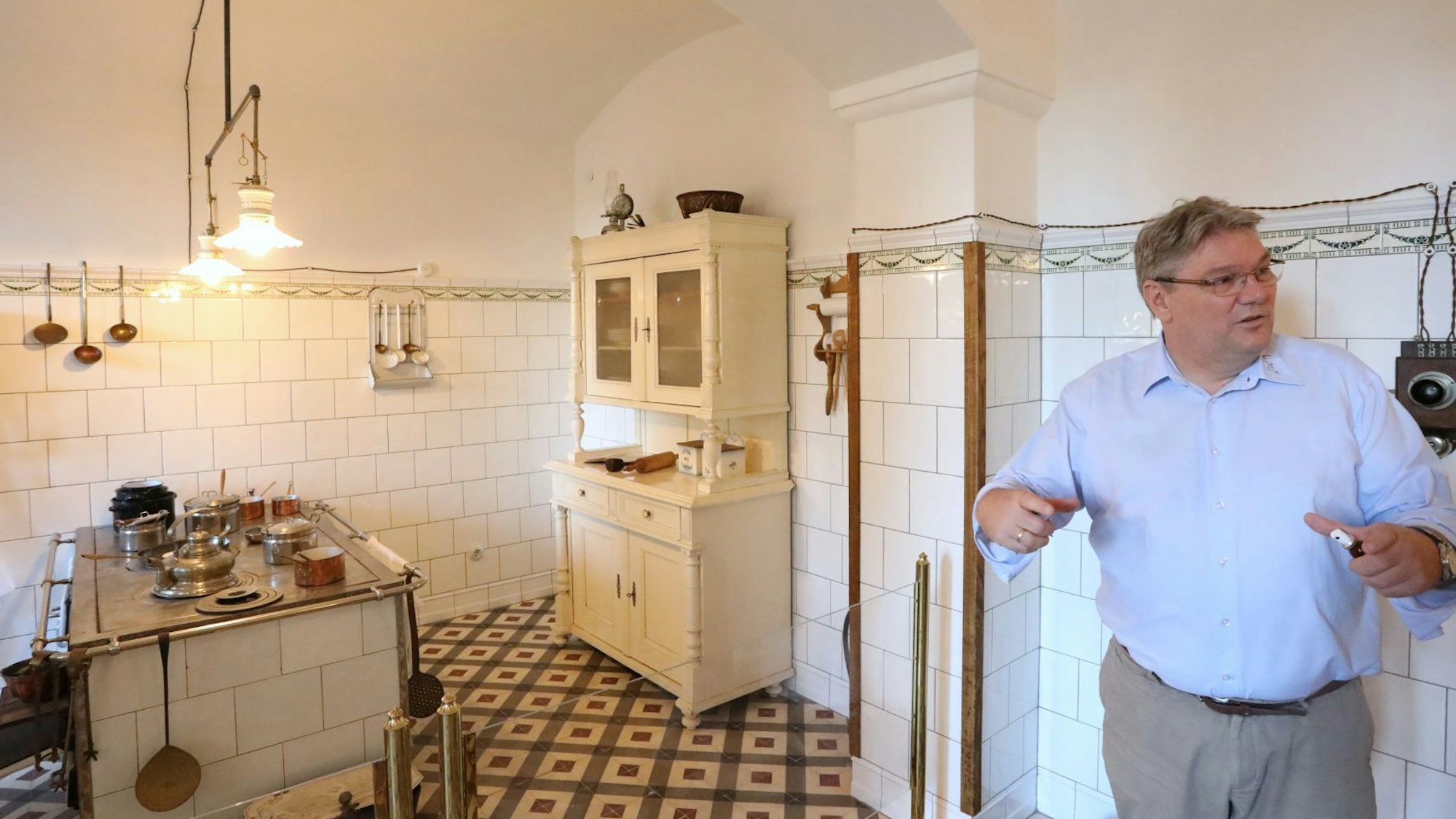 Ein Mann steht in einem Raum mit gefliesten Wänden. In der Küche stehen ein alter Herd und ein alter Küchenschrank,