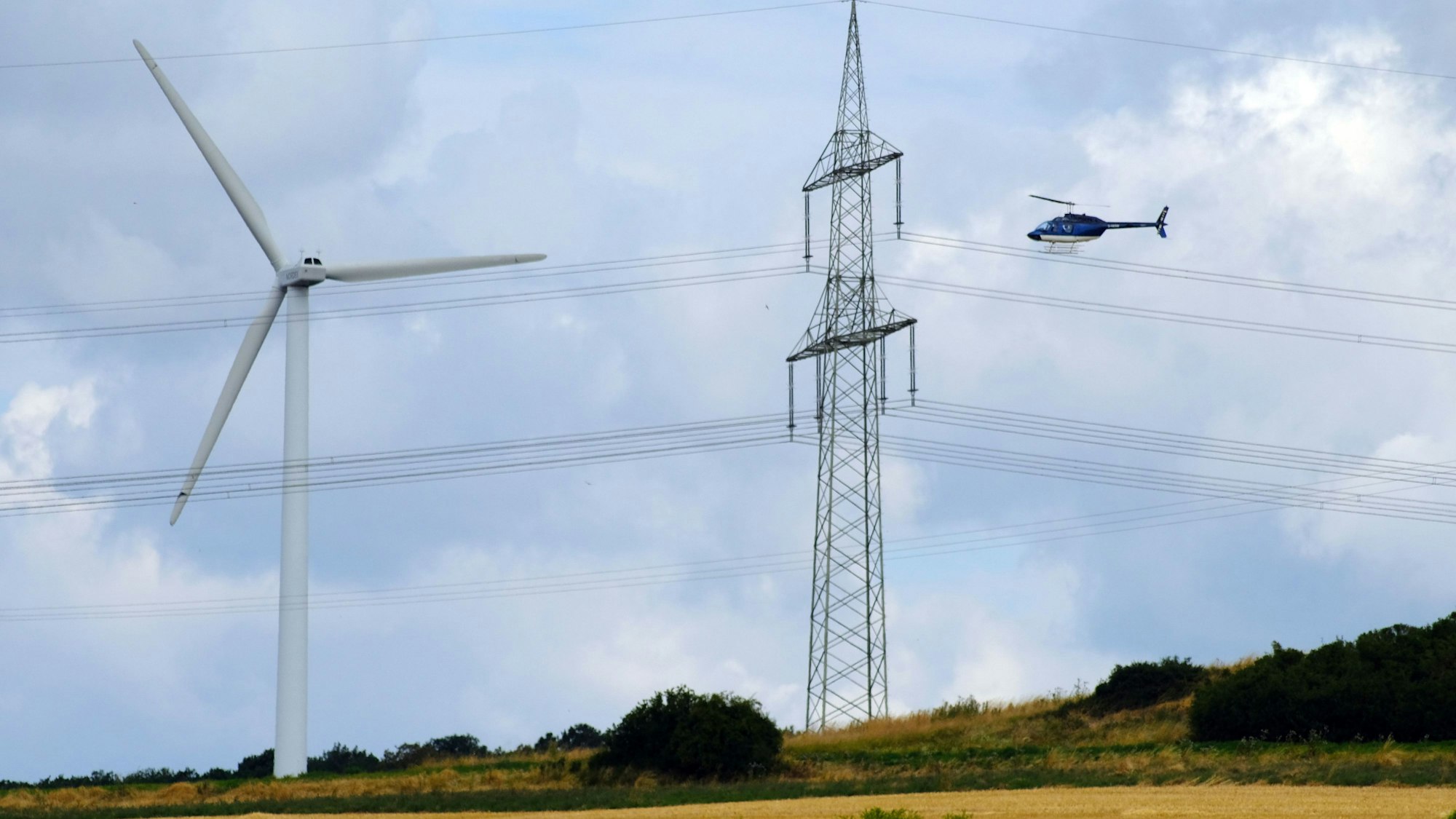 Ein Hubschrauber überfliegt eine 380-KV-Höchstspannungsleitung, links ist eine Windkraftanlage zu sehen.