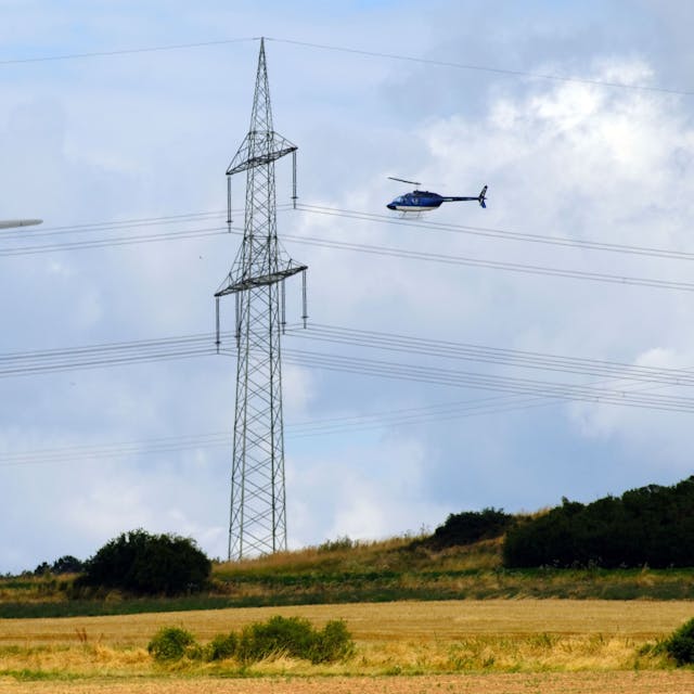 Ein Hubschrauber überfliegt eine 380-KV-Höchstspannungsleitung, links ist eine Windkraftanlage zu sehen.