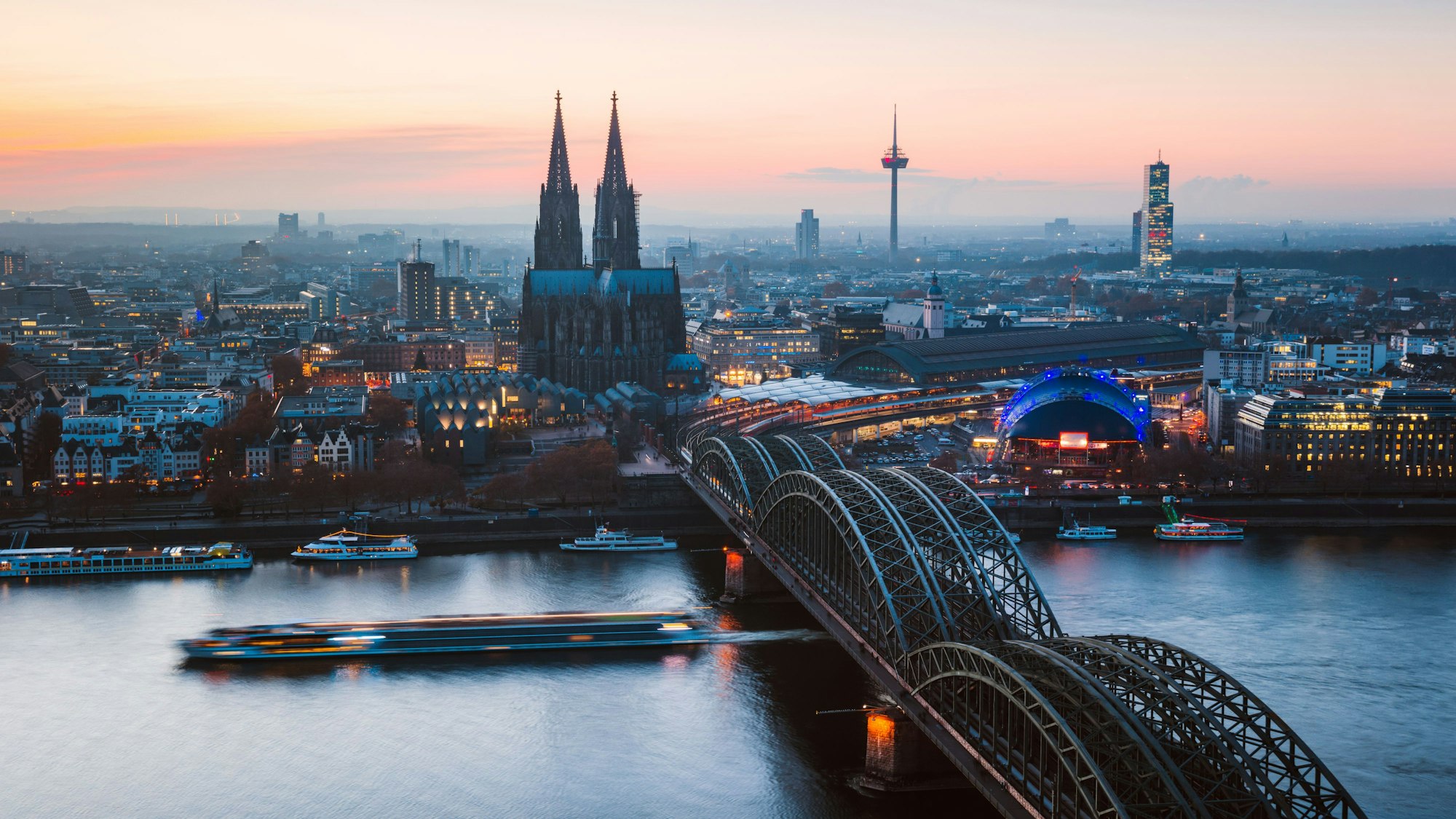 Blick in der Dämmerung über Köln: Im Vordergrund sind die Hohenzollernbrücke und der Kölner Dom zu sehen.