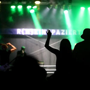 Der Schattenriss von Zuschauern vor der mit grünen Scheinwerfern beleuchteten Bühne mit dem Schriftzug „R(h)einspaziert“.