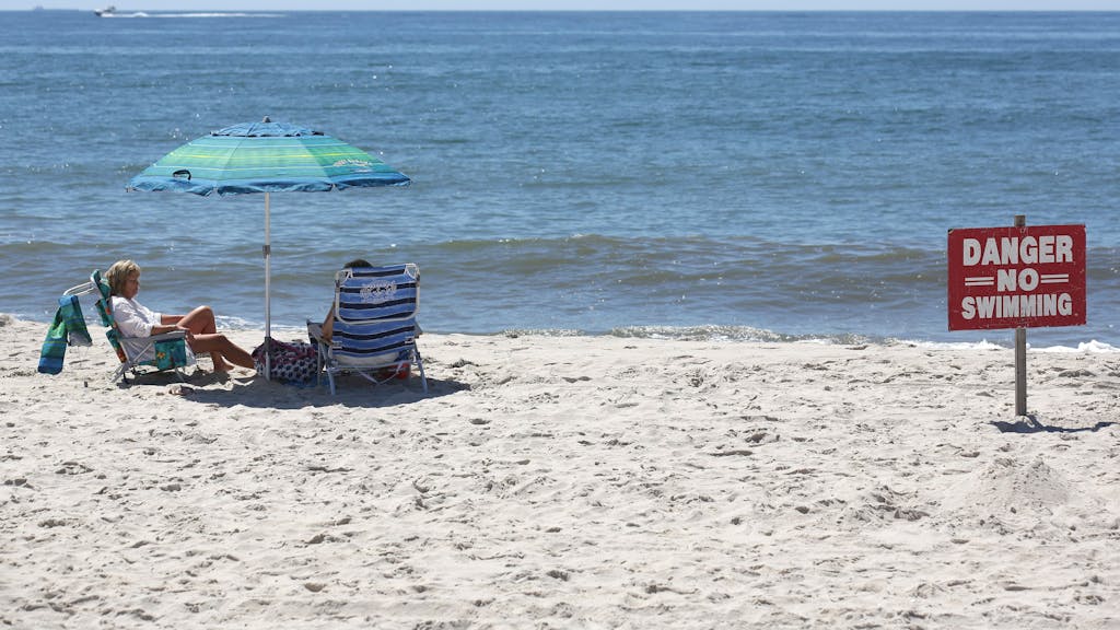 Zwei Menschen sitzen am Strand, neben ihnen steht ein Schild mit der Aufschrift "Vorsicht, nicht schwimmen".