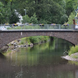 Zu sehen ist die beschädigte Brücke über die Olef in Schleiden. Am Geländer hängen Blumenkübel, links im Bild sind Bäume zu erkennen.
