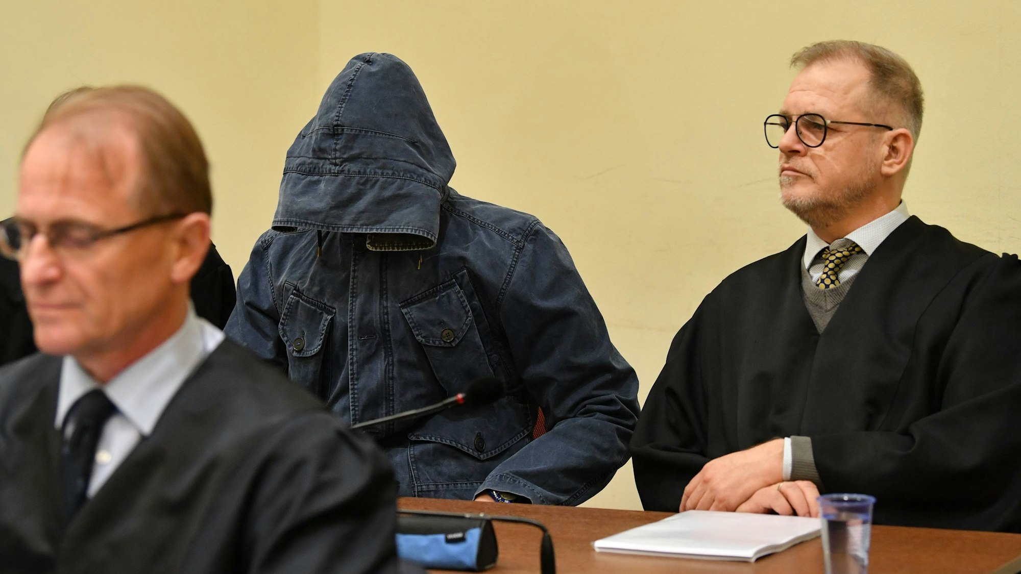 Das Bild zeigt einen Verhandlungstag im NSU-Prozess 2018: Der Angeklagte Carsten S. trägt eine dunkelblaue Jacke, die Kapuze ist so vor sein Gesicht gezogen, dass man es nicht erkennen kann. Rechts neben ihm sitzt sein Anwalt Jacob Hösl hinter einem Tisch im Saal im Landgericht in München.