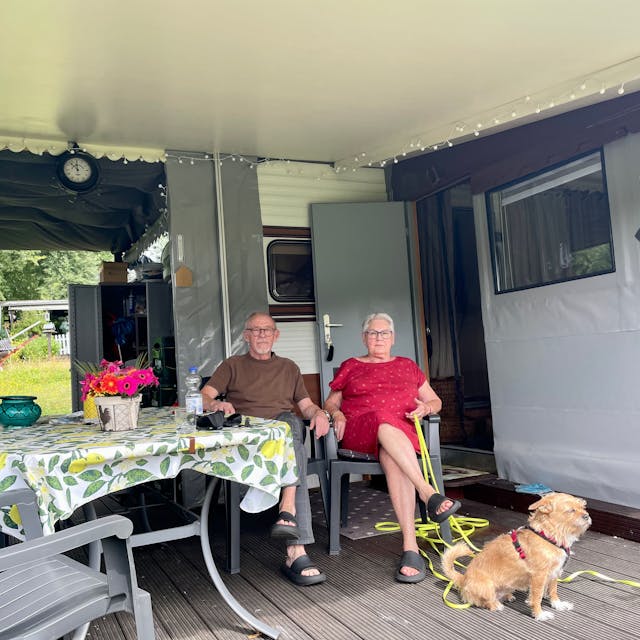 Zwei Senioren mit Hund sitzen in Campingstühlen vor ihrem Dauercamper-Wohnwagen.