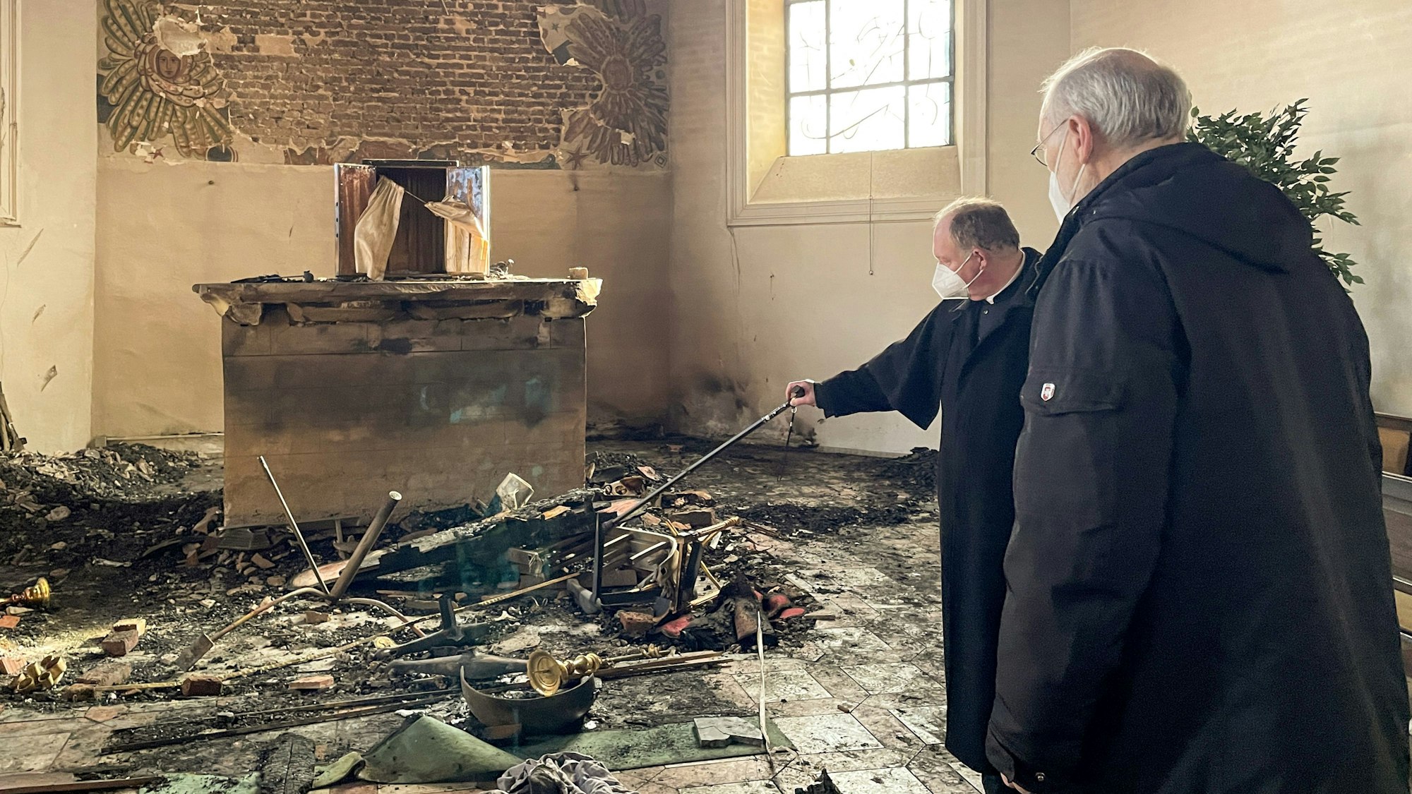 Zwei Priester stehen in einem Kircheninnenraum vor Überrresten nach einem Brand.