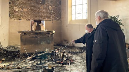 Zwei Priester stehen in einem Kircheninnenraum vor Überrresten nach einem Brand.