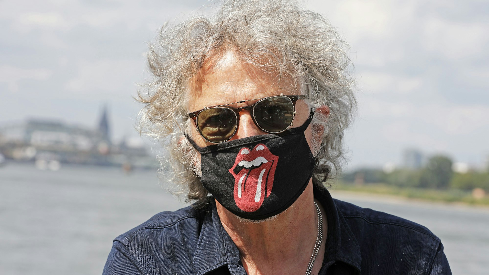 Der Musiker und Sänger Wolfgang Niedecken steht mit einem Mundschutz mit dem Zungen-Logo der Rolling Stones am Rhein.