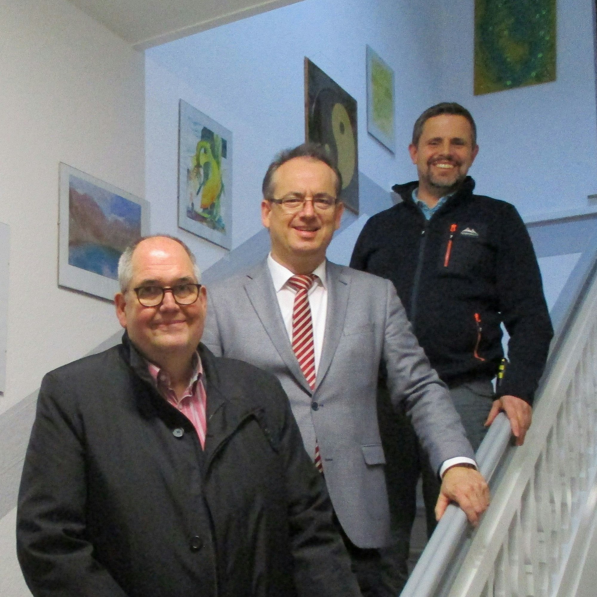 Von der Rheinbacher Umsetzung der NRW-Knastkulturwoche 2022 überzeugte sich der Leitende Ministerialrat Dr. Stephan Neuheuser (Mitte) persönlich. Im Hintergrund sind verschiedenen Malereien zu sehen, zwei Männer in Anzügen sind Neuheusers Begleiter.