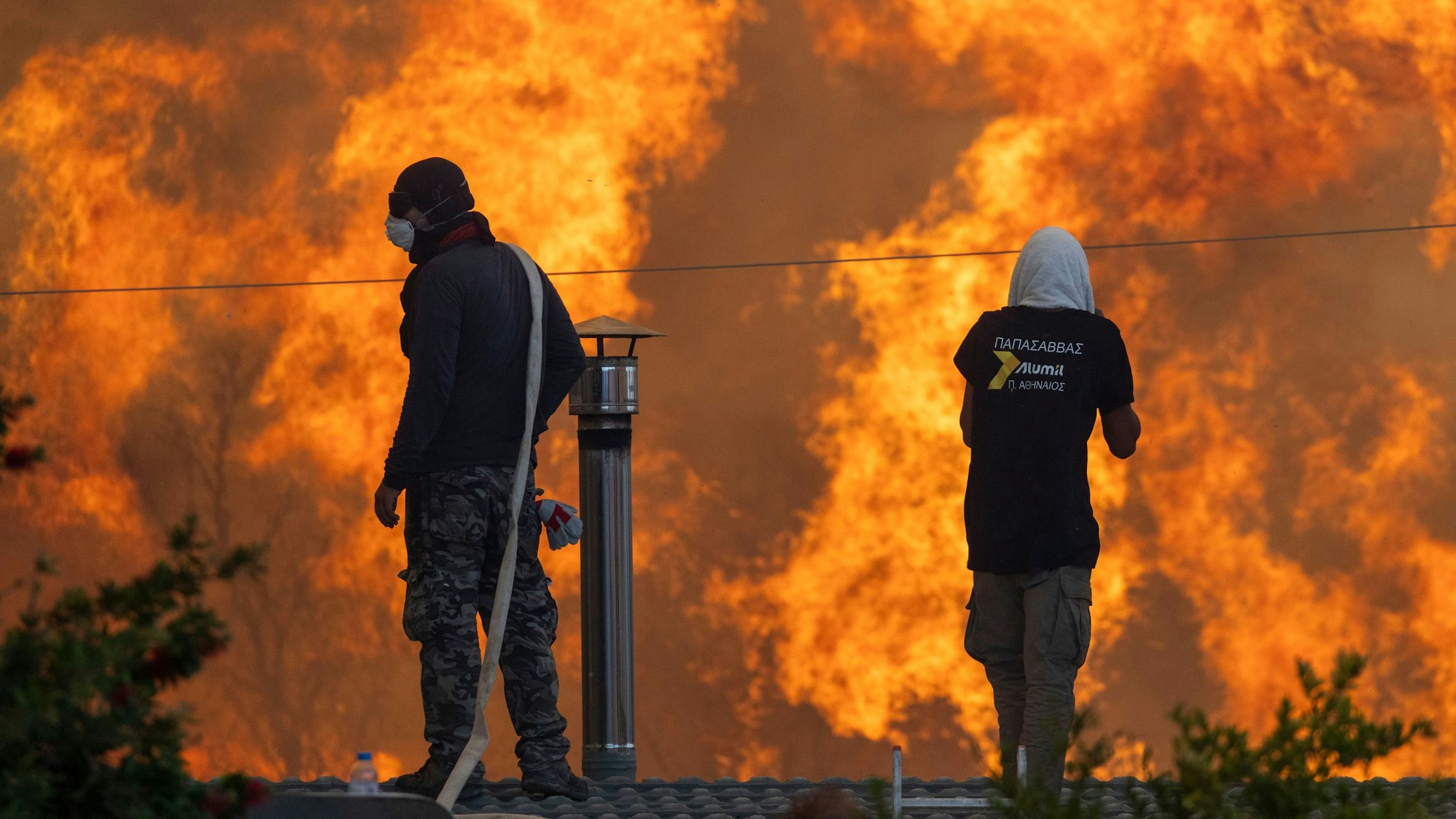 Zwei Männer stehen auf ihren Dächern vor riesigen Flammen und versuchen mit Wasserschläuchen ihre Häuser zu retten.