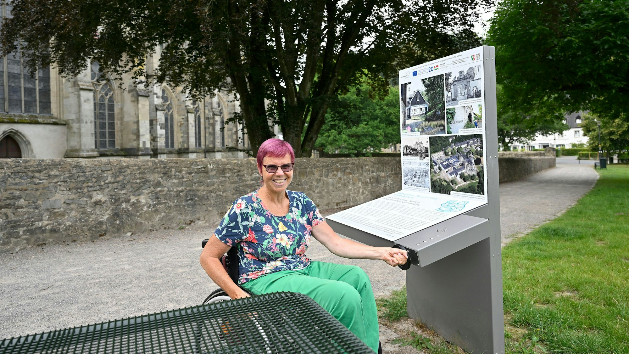 Eine Frau sitzt im Rollstuhl und bedient lachend eine Kurbel, mit der eine Infostation am Altenberger Dom bedient wird.