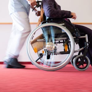 Ein Pfleger schiebt eine Bewohnerin eines Pflegeheims in einem Rollstuhl über den Flur.&nbsp;