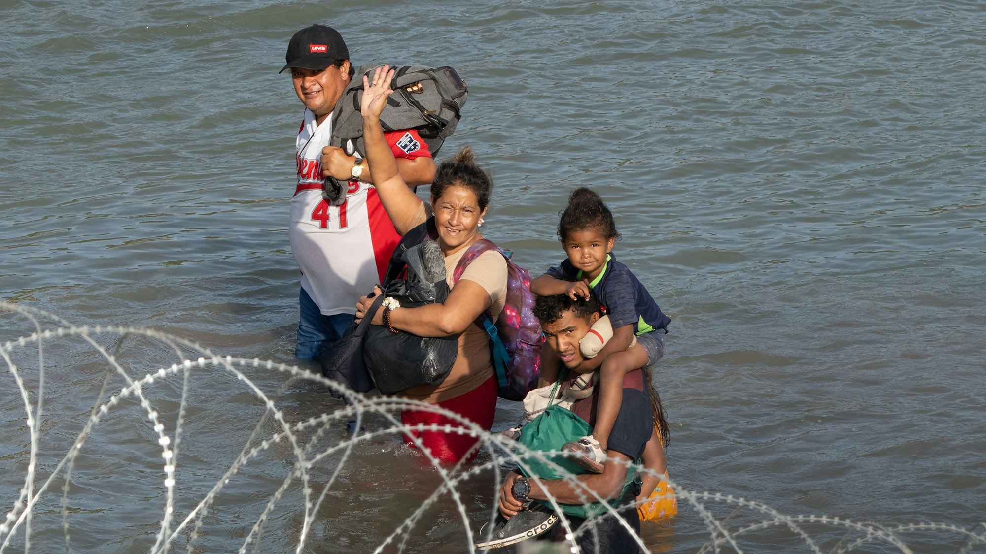 Migranten winken vor Stacheldraht als sie durch den Rio Grande laufen.