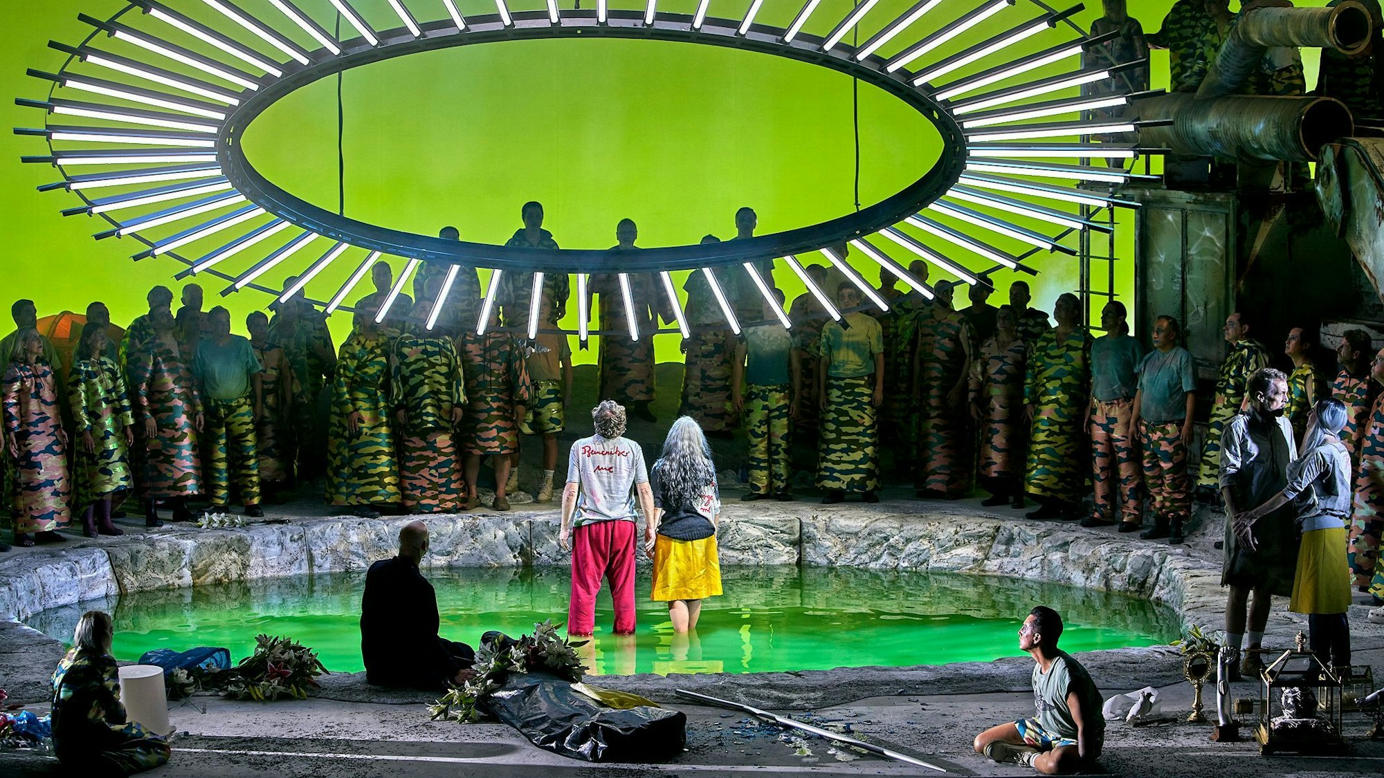 Auf der Bühne stehen Parsifal und Kundry in einer Art Teich, über ihnen ein großer Kreis mit LED-Lampen. Um sie herum stehen zahlreiche Menschen.