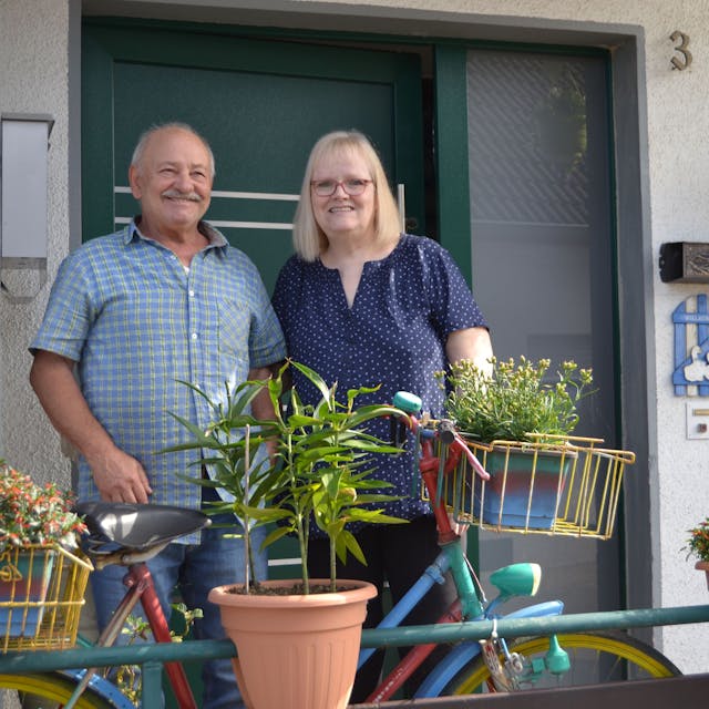 Ein Ehepaar steht vor einer Haustüre. Vor ihnen steht ein bepflanztes Fahrrad.&nbsp;