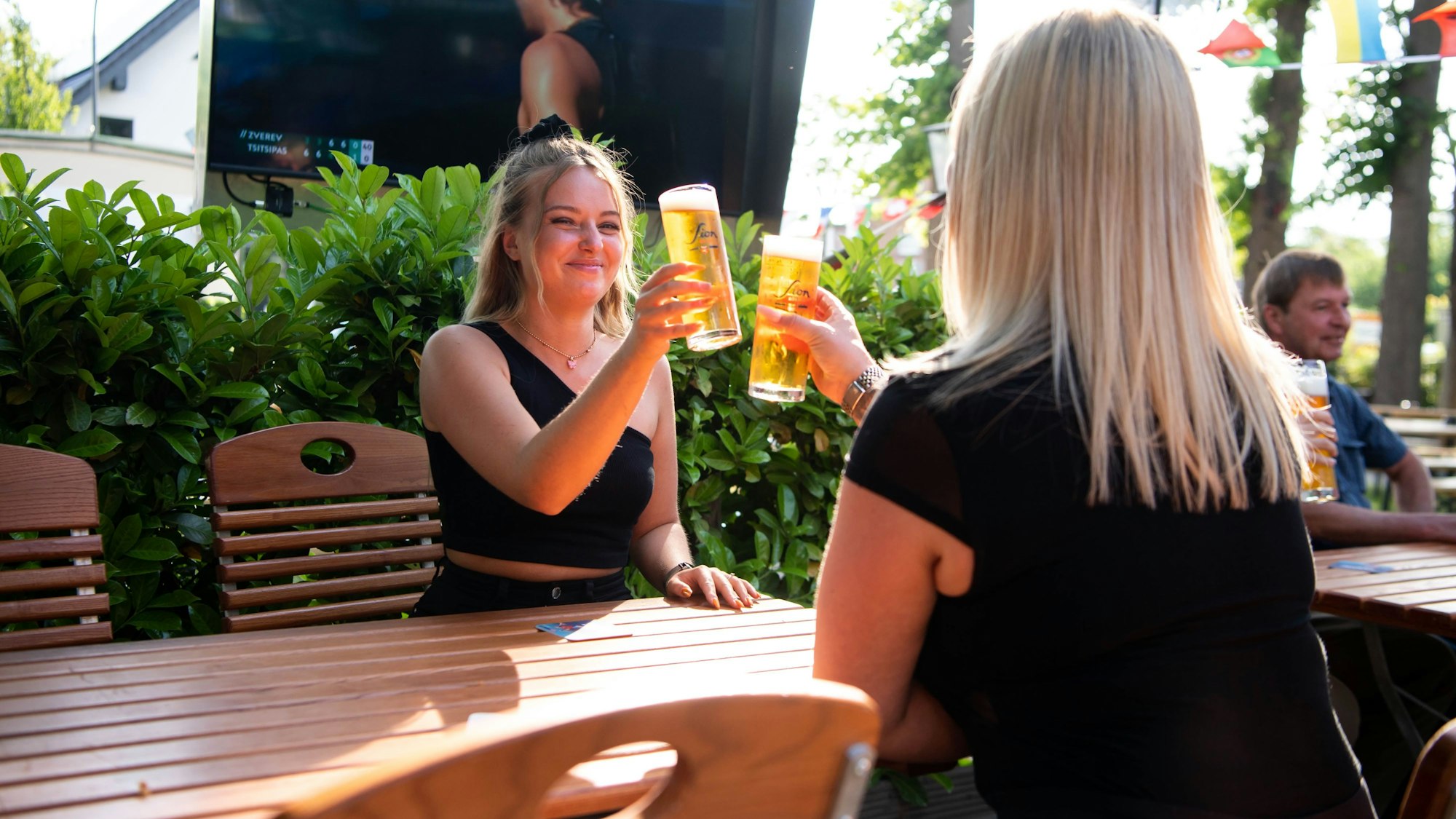 Zwei Frauen sitzen an einem Biergartentisch und stoßen mit einem Bier an.