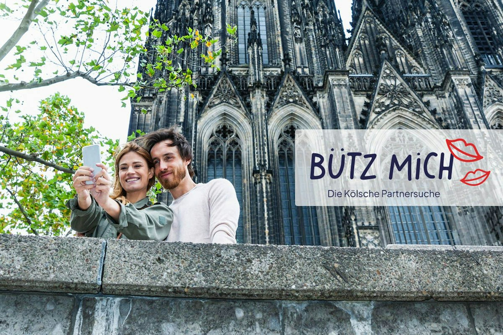Mann und Frau schießen Selfie vorm Kölner Dom. Daneben das Logo der Website Bütz mich.