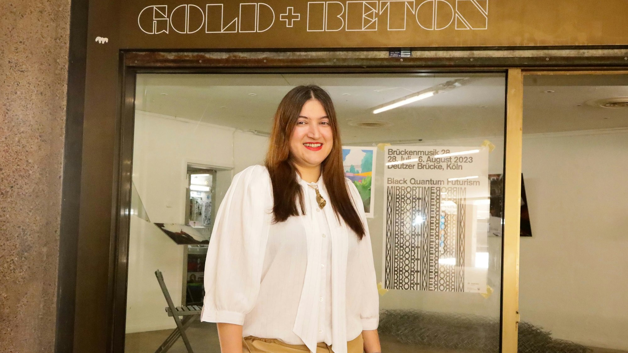 Meryem Erkus steht vor der Galerie „GOLD+BETON". Sie trägt ein weißes Hemd. Hinter ihr bewirbt auf einer Scheibe ein Plakat die Veranstaltung „Brückenmusik“.