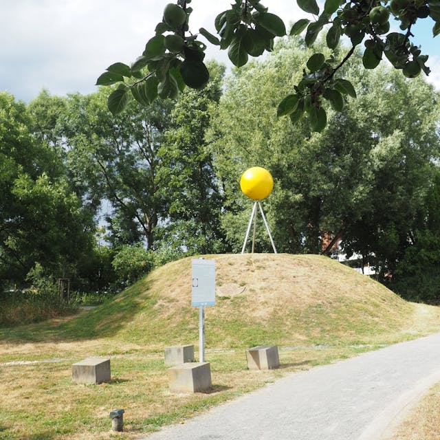 Auf einem kleinen Hügel, der nur spärlich mit Gras bewachsen ist, steht eine große gelbe Kugel auf drei Metallstangen.