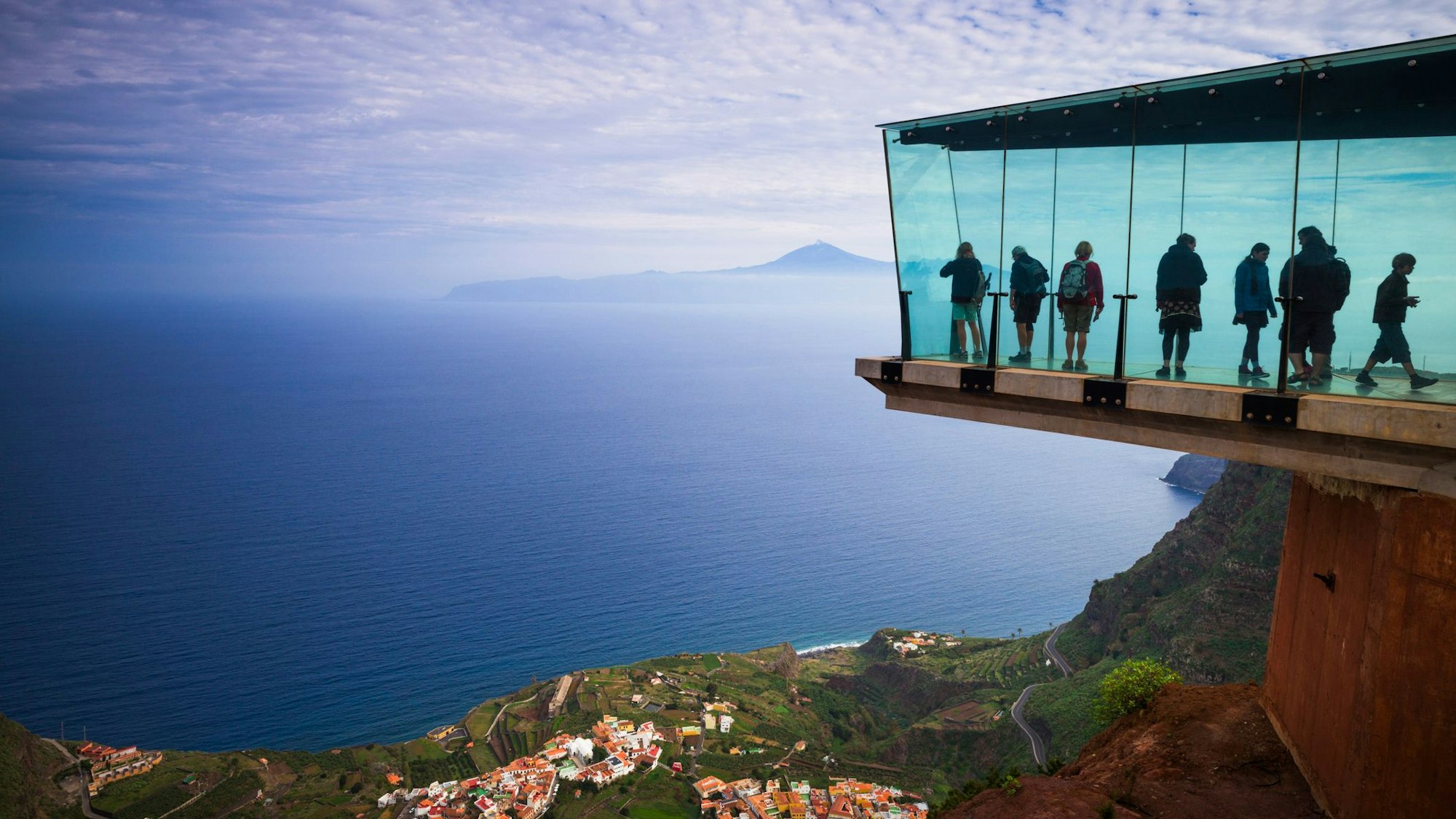 Verschiedene Personen stehen auf einer gläsernen Aussichtsplattform. Unter ihnen ein Tal mit Häusern, im Hintergrund ein Meer mit einer Insel.