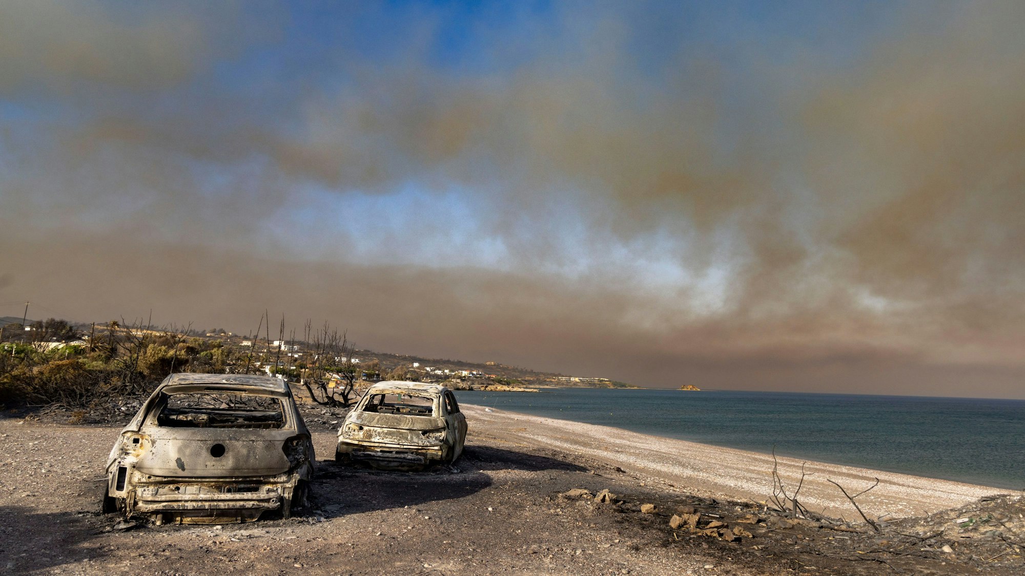 In der Ortschaft Kiotari stehen zwei ausgebrannte PKWs am Strand, am Himmel treibt der Wind eine riesige Rauchwolke in Richtung Nordosten.