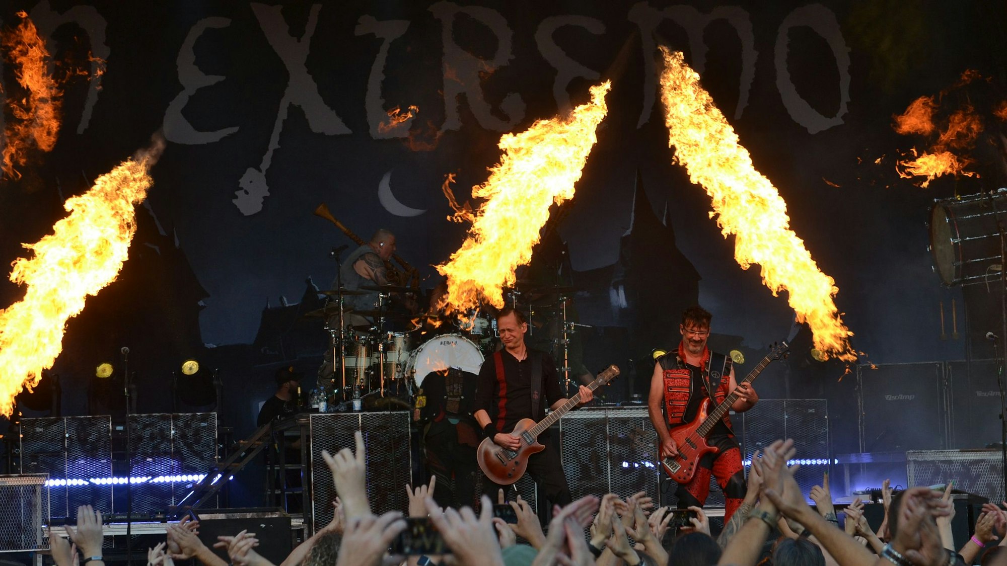 Eine Band steht auf einer Bühne, zur Show gehören auch Feuer-Fontänen. Im Vordergrund recken zahlreiche Fans die Hände in die Höhe.