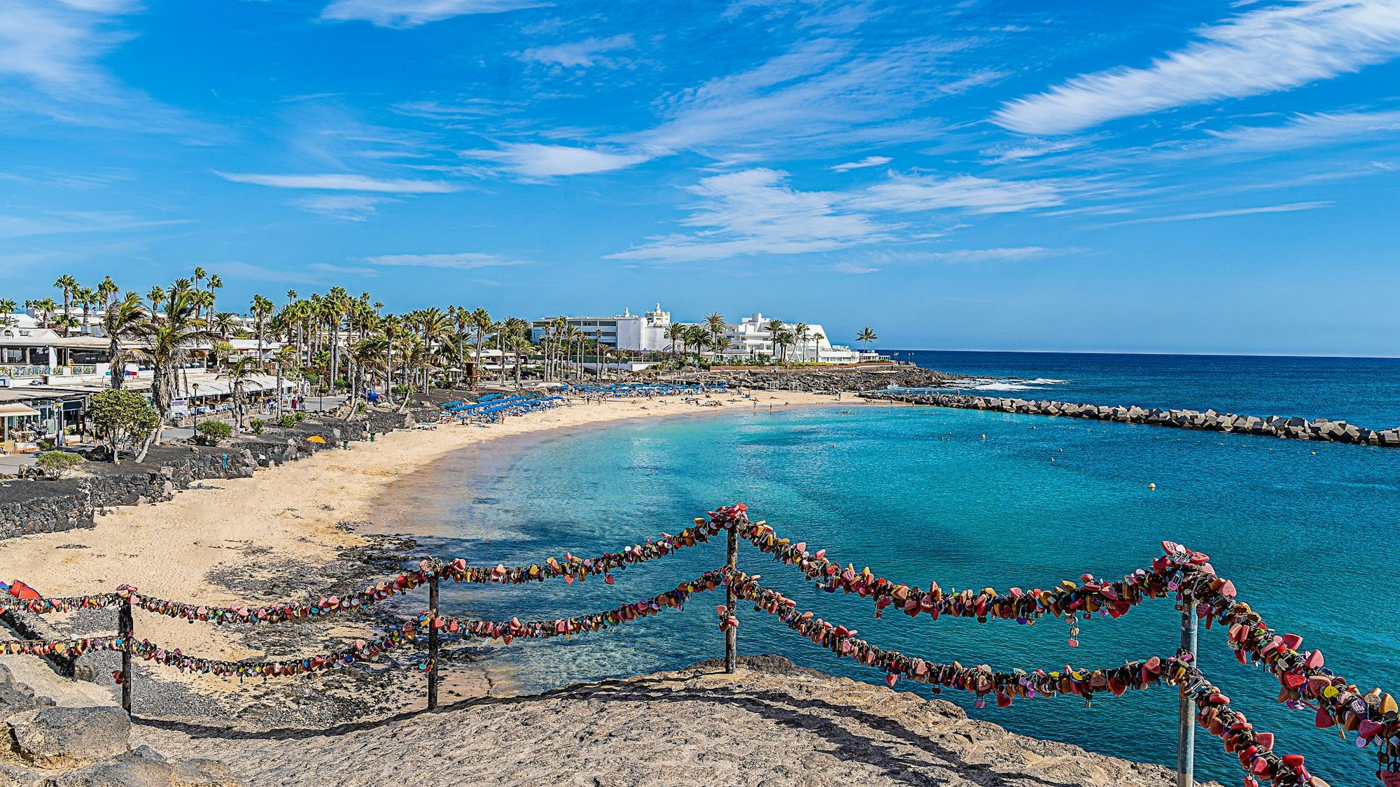 Im Vordergrund ist ein Geländer mit Schlössern zu sehen. Im Hintergrund grenzt ein Strand mit Palmen und einer Promenade an blaues Meer.