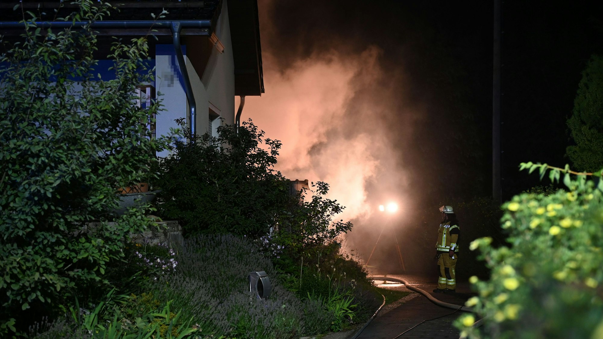 Das Bild zeigt einen Teil eines Wohnhauses, dahinter steigt Rauch auf, ein Feuerwehrmann betrachtet die Szenerie.