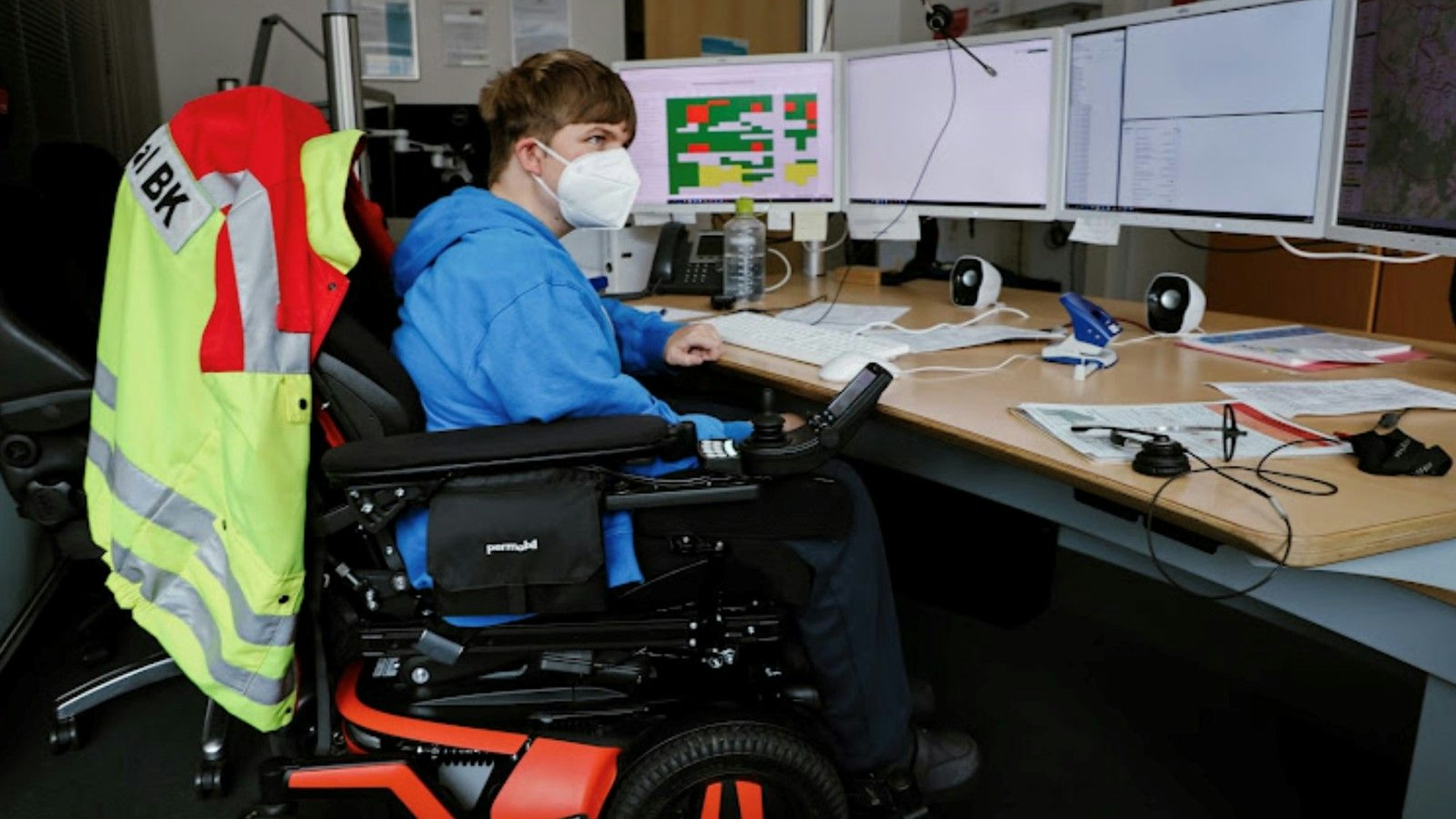 Ein junger Mann mit Maske sitzt in einem Rollstuhl vor einem Schreibtisch. Er schaut konzentriert auf die vier Computer-Bildschirme vor sich.