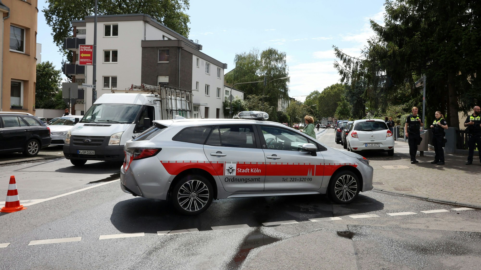 Ein Auto mit der Aufschrift Stadt Köln Ordnungsamt steht quer auf der Straße, rechts sind Mitarbeitende des Ordnungsamtes zu sehen.