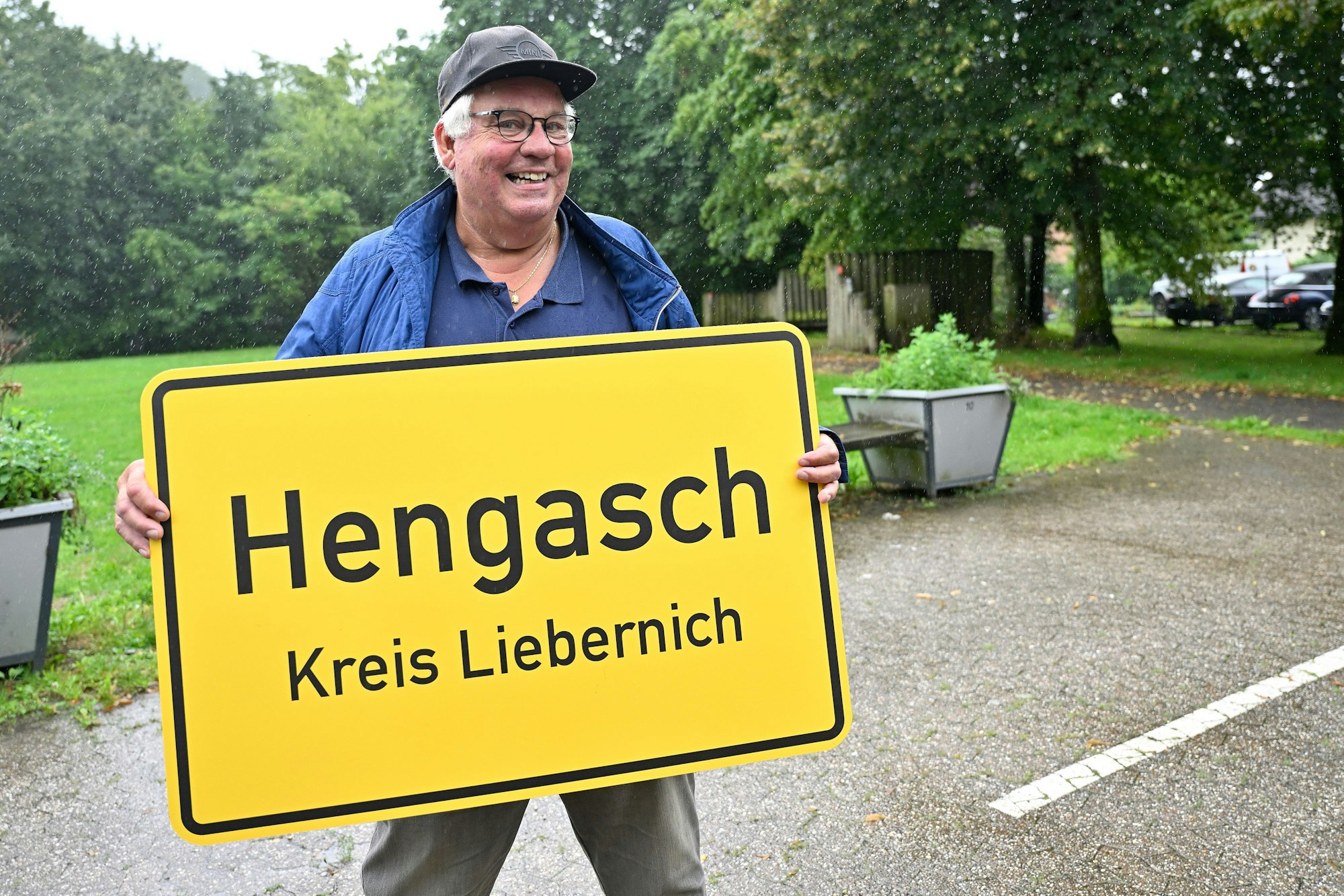 Ein Mann hält eine gelbe Ortstafel in Händen, auf dieser steht: Hengasch Kreis Liebernich.