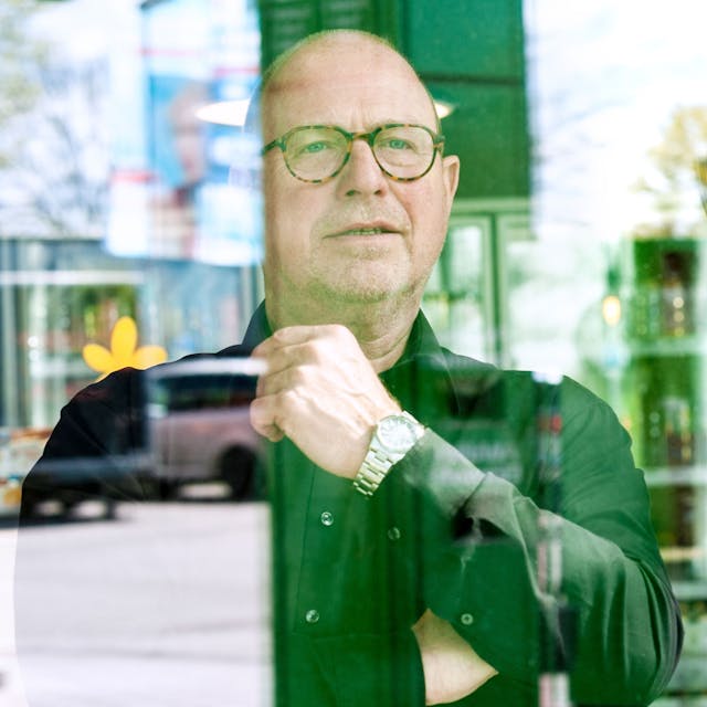 Karl-Heinz Breuer schaut aus dem Schaufenster, in dessen Spiegelung Teile der Tankstelle zu sehen sind. Hinter ihm stehen Getränke.