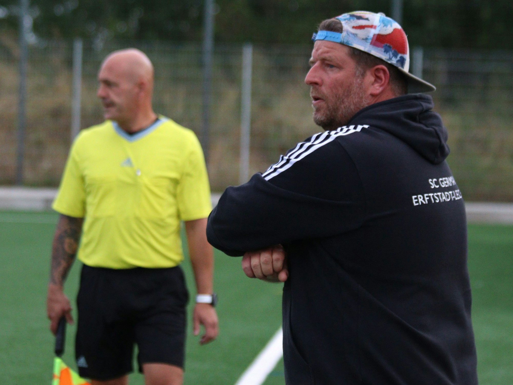 Karsten Kochems, Trainer von Erftstadt-Lechenich, trägt eine bunte Kappe verkehrt herum auf dem Kopf. Im Hintergrund Linienrichter Jörg Piana.