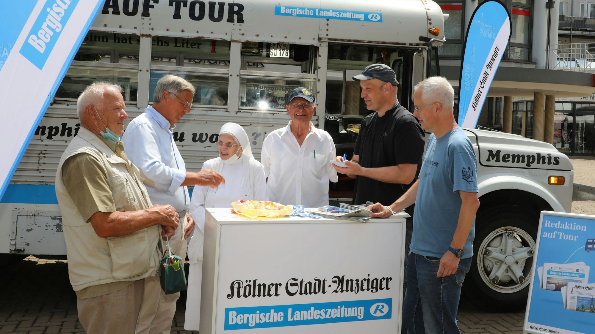 Menschen stehen um eine Infotheke vor einem historischen amerikanischen Schulbus. Auf der Theke und auf Fahnen sind die Zeitungstitel Bergische Landeszeitung und Kölner Stadt-Anzeiger zu lesen.