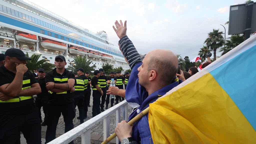 Ein Mann mit einer ukrainischen Flagge steht vor einer Reihe an Polizisten. In Hintergrund ist das russische Kreuzfahrtschiff „Astoria Grande“ zu sehen.