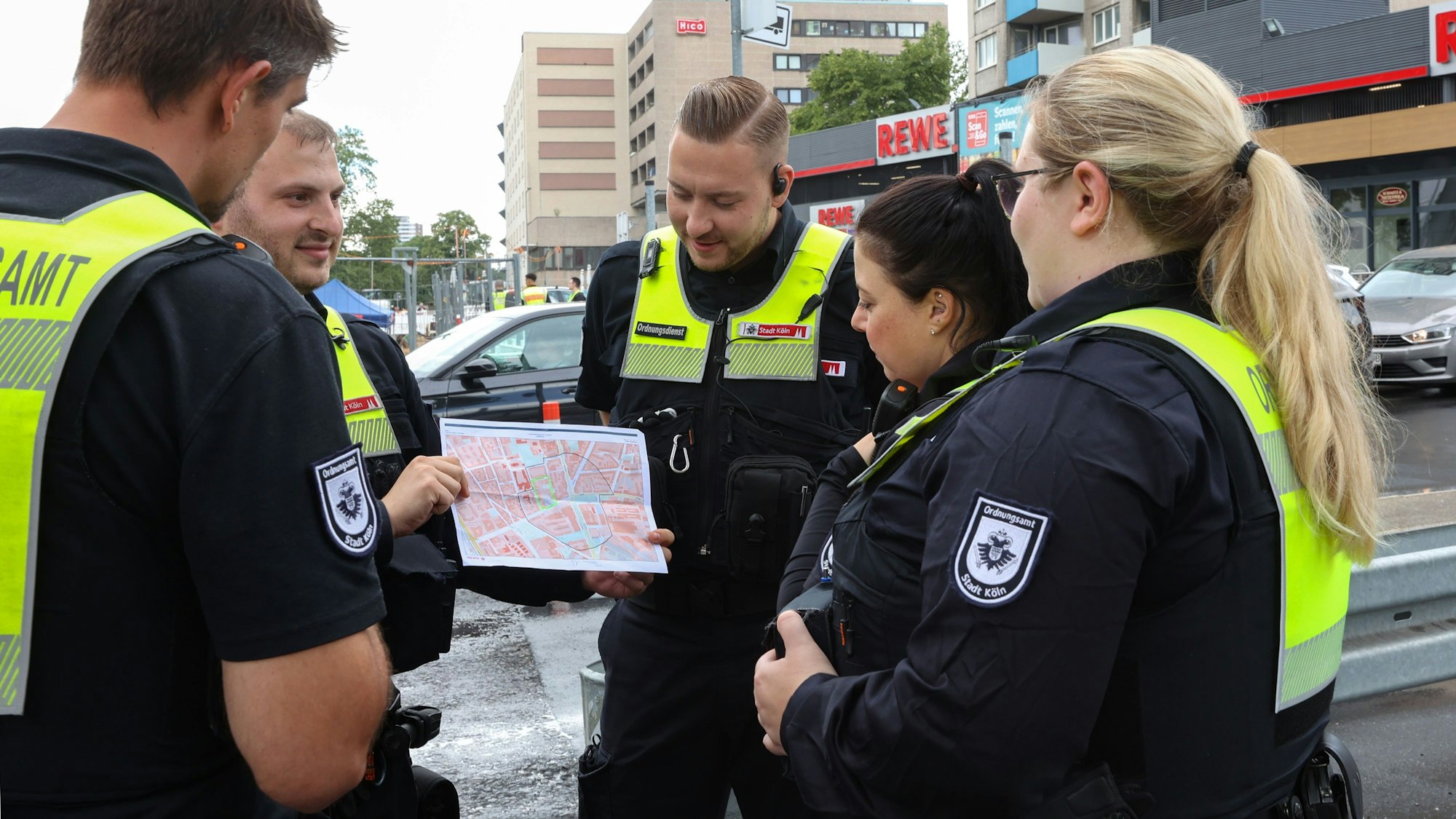 Mitarbeitende in Dienstkleidung der Stadt Köln stehen im Kreis, ein Mitarbeiter hält eine ausgedruckte Karte mit einem Stadtplan in der Hand.