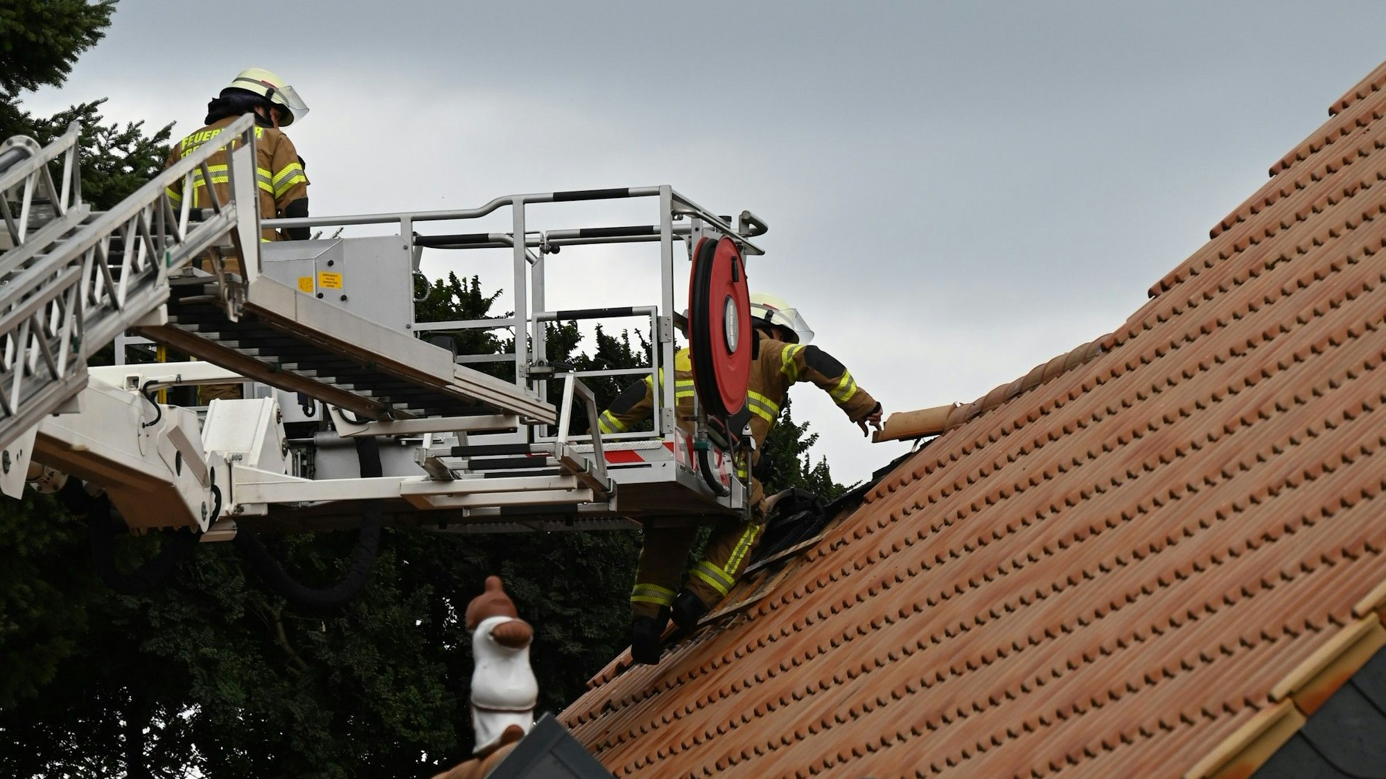 Das Bild zeigt Feuerwehrleute an einem Hausdach, die die Ziegel entfernen.